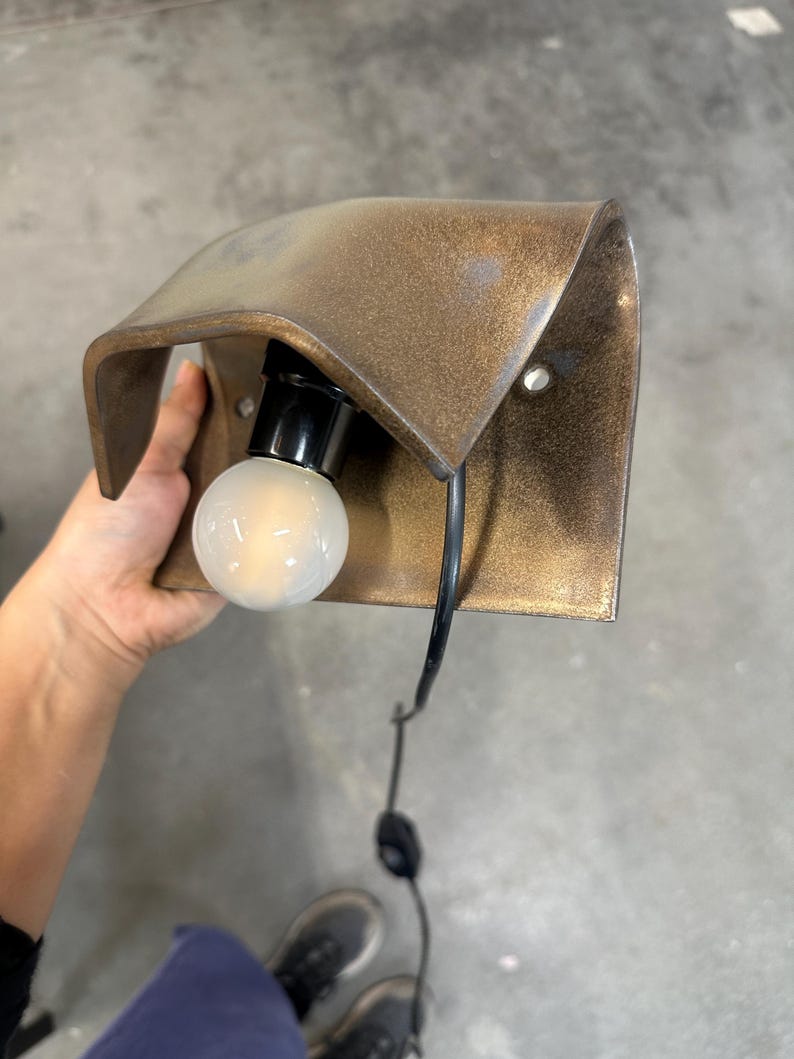 Pode incluir: Um candeeiro de parede com um abajur de metal curvo em tom de bronze com acabamento texturizado. A lumin&aacute;ria inclui um casquilho preto com uma l&acirc;mpada redonda de vidro fosco branco e um cabo de alimenta&ccedil;&atilde;o preto com interruptor.