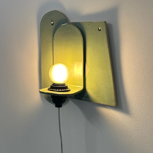 May include: A wall-mounted light fixture featuring a pale green, speckled ceramic backplate with an arched design. A small shelf extends from the center, holding a glowing, round light bulb in a black socket with a thin white power cord hanging below.