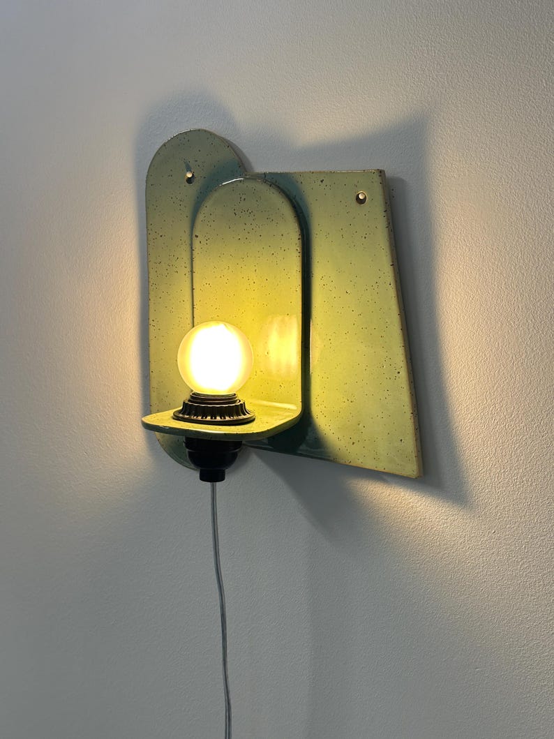 May include: A wall-mounted light fixture featuring a pale green, speckled ceramic backplate with an arched design. A small shelf extends from the center, holding a glowing, round light bulb in a black socket with a thin white power cord hanging below.