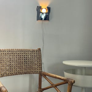 May include: A wall-mounted lamp with a glossy, dark metallic finish and a curved, sculptural form. The lamp emits a warm, glowing light from a central bulb. A thin white power cord hangs vertically from the base of the fixture down the wall.