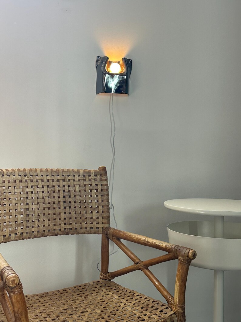May include: A wall-mounted lamp with a glossy, dark metallic finish and a curved, sculptural form. The lamp emits a warm, glowing light from a central bulb. A thin white power cord hangs vertically from the base of the fixture down the wall.