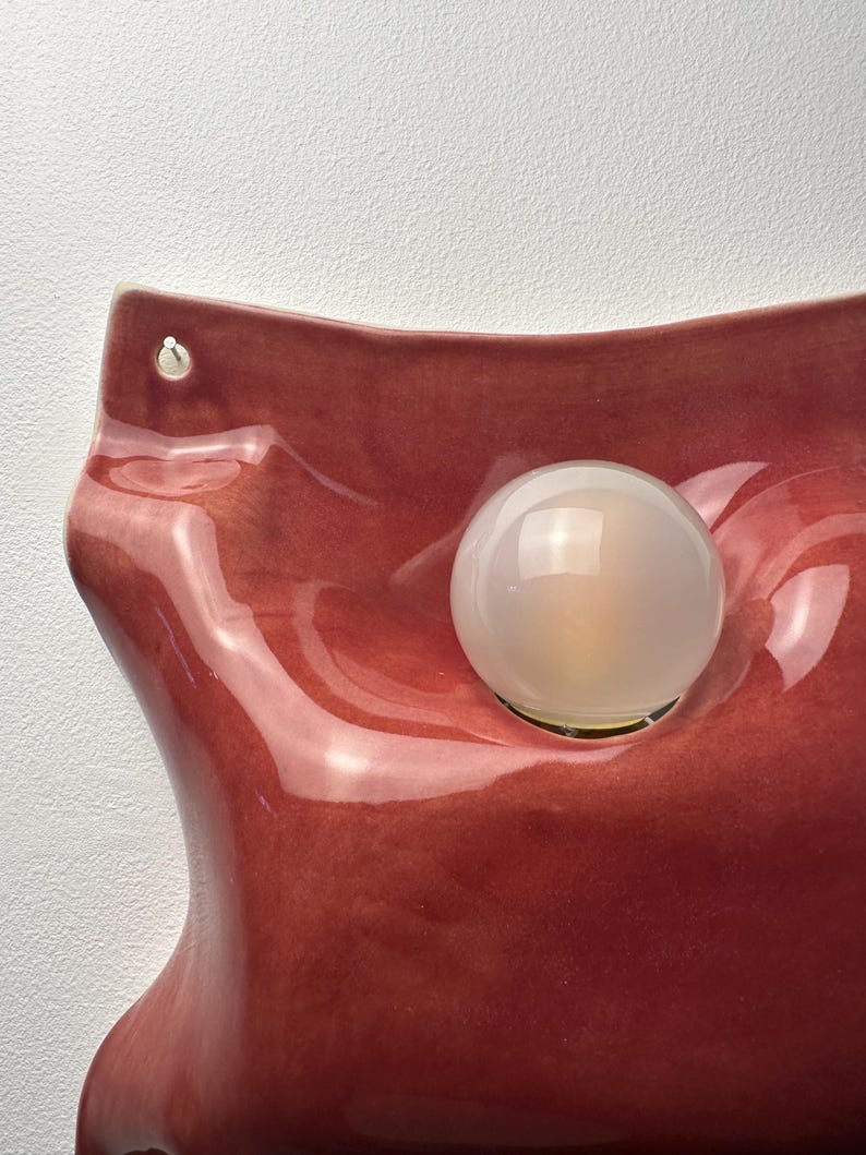K&ouml;nnte beinhalten: Eine Wandleuchte mit einem gl&auml;nzenden, dunkelroten Keramiksockel in organischer, gewellter Form. Eine einzelne, kugelf&ouml;rmige, mattwei&szlig;e Glasgl&uuml;hbirne ist in eine vertiefte, kreisf&ouml;rmige &Ouml;ffnung in der Mitte der Keramikform eingelassen.