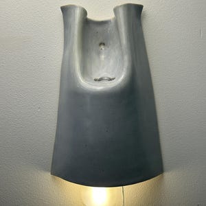 K&ouml;nnte beinhalten: Eine Wandleuchte mit einem glatten, hellgrauen Keramikschirm, der einen geschwungenen, U-f&ouml;rmigen oberen Teil und eine breite, ausgestellte Basis aufweist. Eine einzelne runde, mattierte Gl&uuml;hbirne ist unten sichtbar und strahlt warmes Licht aus.