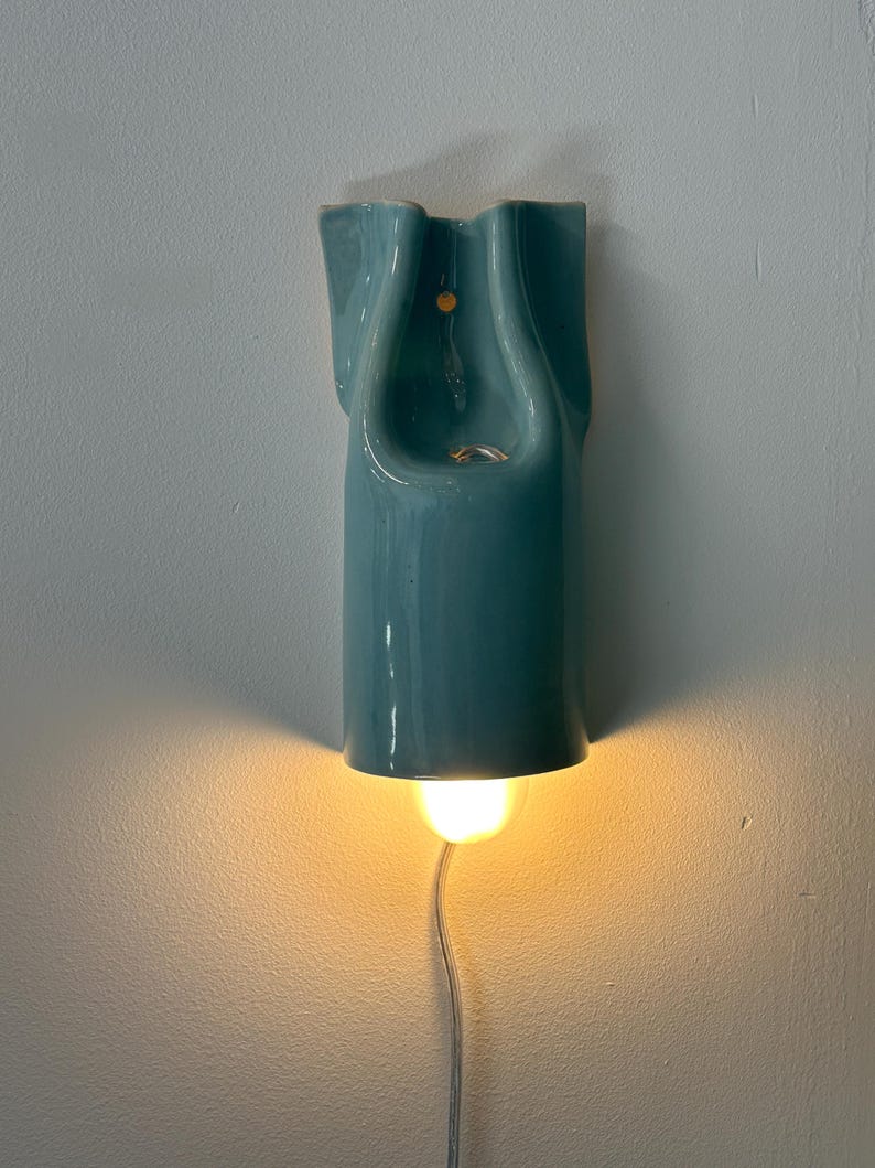 May include: A wall-mounted ceramic light fixture with a glossy teal finish and a sculptural, folded form. The fixture features a central opening and emits a warm, downward-facing glow from a light bulb positioned at the base, with a thin power cord.