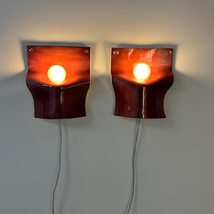 May include: A pair of wall-mounted sconces featuring a curved, glossy red ceramic body with a gradient finish. Each fixture holds a single round, glowing light bulb centered in the middle, with a thin gray power cord extending downward from the base.