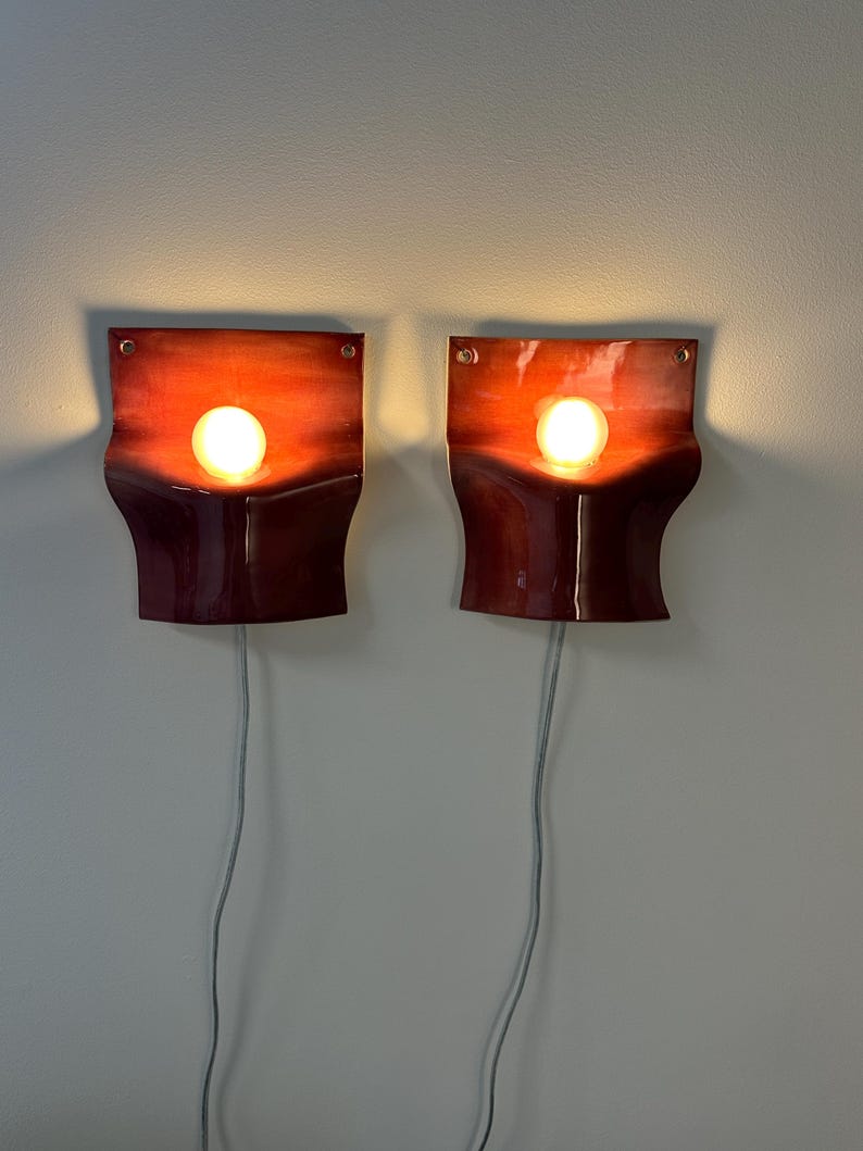 May include: A pair of wall-mounted sconces featuring a curved, glossy red ceramic body with a gradient finish. Each fixture holds a single round, glowing light bulb centered in the middle, with a thin gray power cord extending downward from the base.