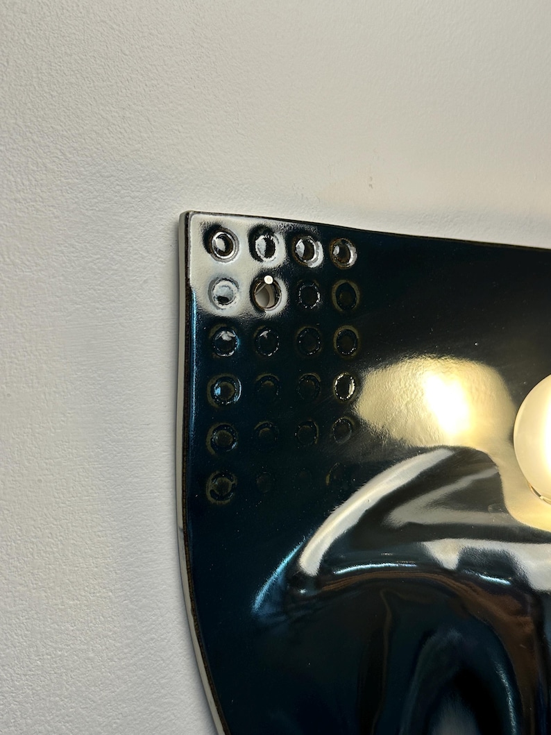 K&ouml;nnte beinhalten: Eine gl&auml;nzende, dunkelblaue Wandleuchte aus Keramik mit einem Gitter aus kreisf&ouml;rmigen Vertiefungen in der oberen Ecke. Die Leuchte hat eine glatte, reflektierende Oberfl&auml;che und eine runde Gl&uuml;hbirne an der rechten Seite.