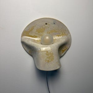 May include: A wall-mounted ceramic lamp with a rounded, organic shape and a textured, mottled cream and yellow glaze. The fixture features a central circular opening holding a single round white light bulb and a thin power cord extending downward.