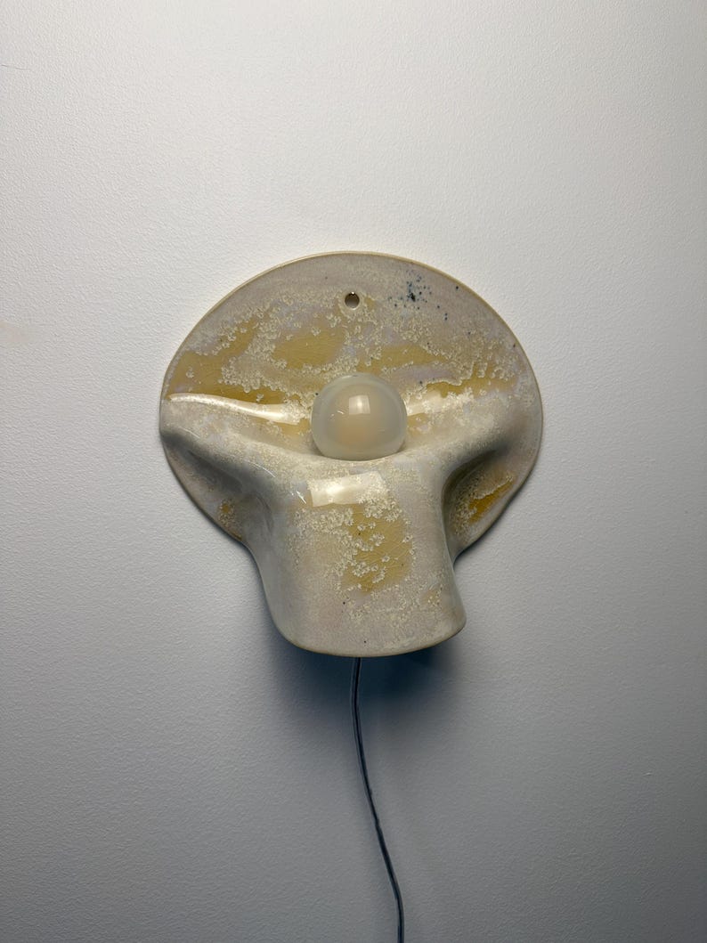 May include: A wall-mounted ceramic lamp with a rounded, organic shape and a textured, mottled cream and yellow glaze. The fixture features a central circular opening holding a single round white light bulb and a thin power cord extending downward.