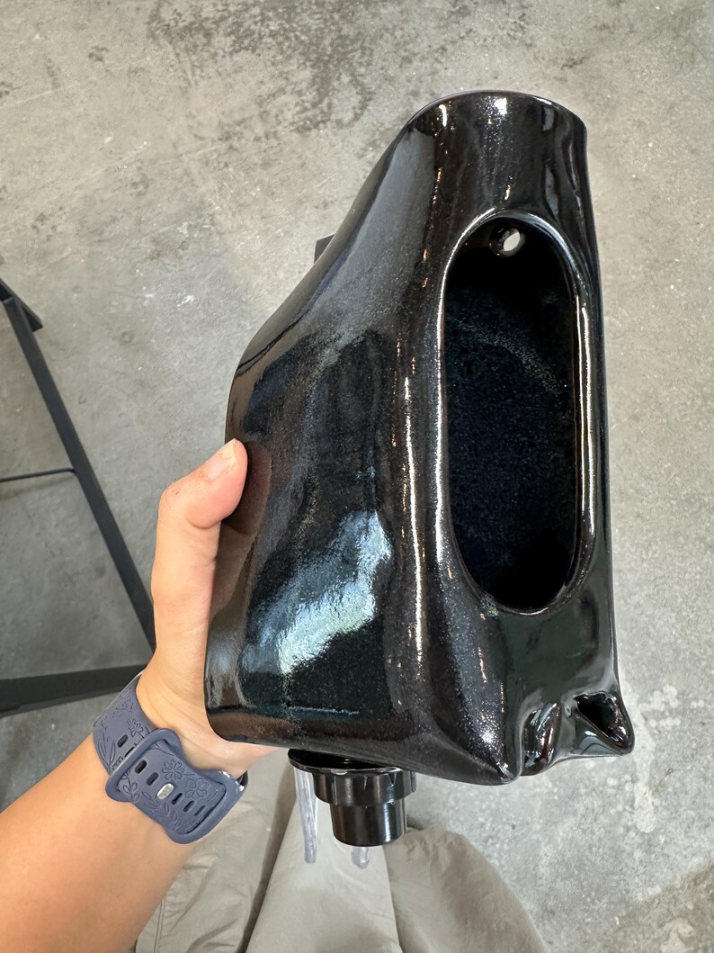 May include: A glossy black ceramic wall-mounted planter with a smooth, curved, and elongated form. The vessel features a large, oval-shaped opening on the front and a small circular hole near the top for mounting, with a black nozzle at the base.