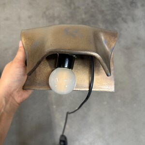 Pode incluir: Um candeeiro de parede com um abajur met&aacute;lico curvo em tom de bronze e uma l&acirc;mpada globo branca central. O candeeiro possui um casquilho preto e um cabo de alimenta&ccedil;&atilde;o preto com um interruptor integrado que sai da base da l&acirc;mpada.