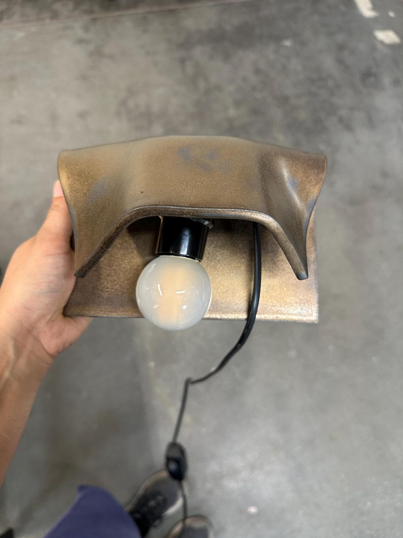 Pode incluir: Um candeeiro de parede com um abajur met&aacute;lico curvo em tom de bronze e uma l&acirc;mpada globo branca central. O candeeiro possui um casquilho preto e um cabo de alimenta&ccedil;&atilde;o preto com um interruptor integrado que sai da base da l&acirc;mpada.