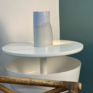 K&ouml;nnte beinhalten: Eine zylindrische Keramik-Tischleuchte mit einer glatten, hellblau-wei&szlig;en Farbverlaufglasur. Die Lampe strahlt ein sanftes, warmes Licht von ihrem Sockel aus und beleuchtet die darunter liegende Fl&auml;che. Sie hat eine schlichte, vertikale Form.