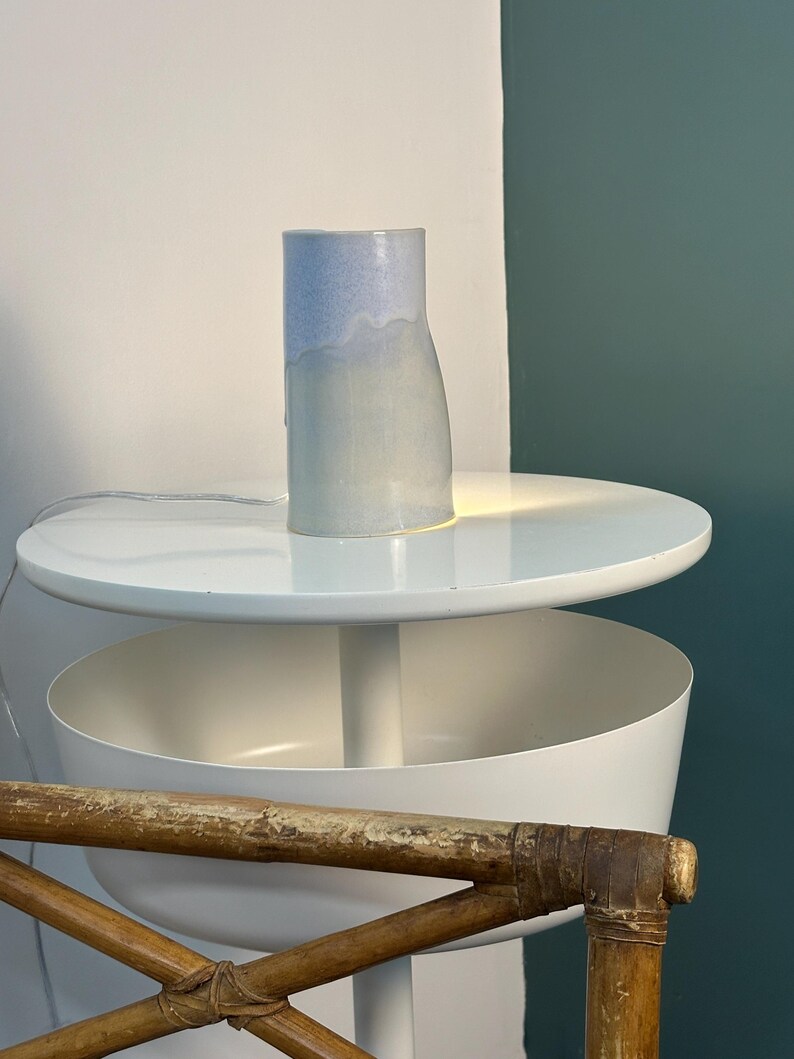 K&ouml;nnte beinhalten: Eine zylindrische Keramik-Tischleuchte mit einer glatten, hellblau-wei&szlig;en Farbverlaufglasur. Die Lampe strahlt ein sanftes, warmes Licht von ihrem Sockel aus und beleuchtet die darunter liegende Fl&auml;che. Sie hat eine schlichte, vertikale Form.
