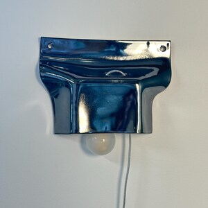 May include: A wall-mounted lamp featuring a glossy, dark blue, metallic-finished ceramic shade with a folded, fluid shape. The fixture includes two circular mounting holes at the top, a central loop, and a single round, frosted light bulb.