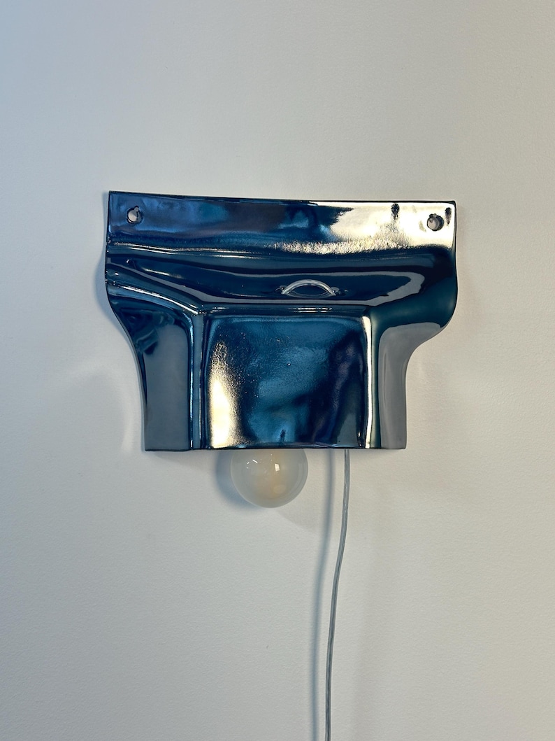 May include: A wall-mounted lamp featuring a glossy, dark blue, metallic-finished ceramic shade with a folded, fluid shape. The fixture includes two circular mounting holes at the top, a central loop, and a single round, frosted light bulb.
