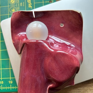 以下が含まれることがあります： 光沢のある深いマゼンタ色の釉薬が施された、有機的で折り重なった形状のセラミック製ウォールランプ。中央のくぼみに配置された円形の白いガラス電球と、取り付け用の2つの小さな円形の穴が特徴です。