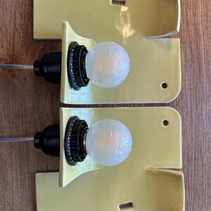 May include: Two wall sconces featuring curved, glossy pale yellow ceramic backplates. Each fixture includes a black socket holding a round, frosted white light bulb. The ceramic plates have rectangular and circular cutouts for mounting purposes.