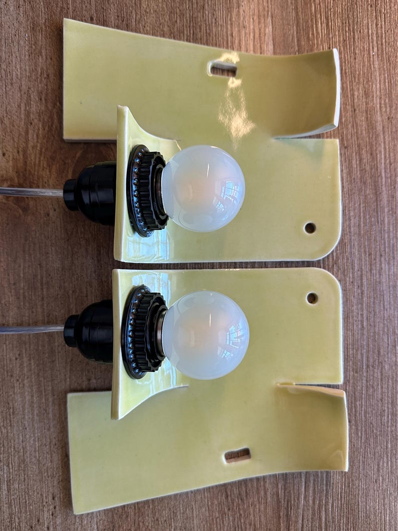 May include: Two wall sconces featuring curved, glossy pale yellow ceramic backplates. Each fixture includes a black socket holding a round, frosted white light bulb. The ceramic plates have rectangular and circular cutouts for mounting purposes.