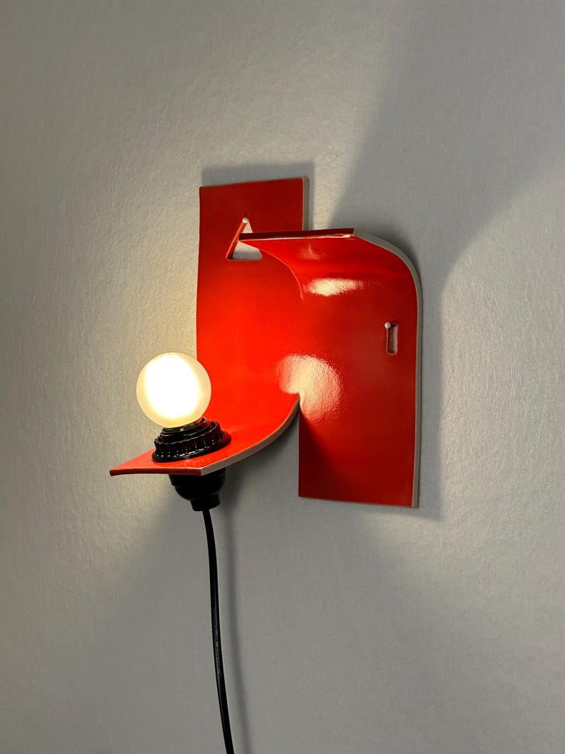 May include: A contemporary, red wall light with a distinctive, geometric shape. It has a small, round light bulb and a black power cable. The light is mounted on a small shelf, and is attached to the wall.