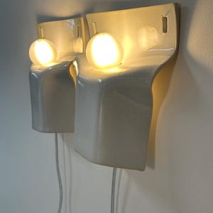 May include: A pair of off-white ceramic wall sconces with a curved, sculptural silhouette. Each fixture features a single glowing spherical light bulb positioned in a recessed top section, with a thin power cord extending downward from the base.