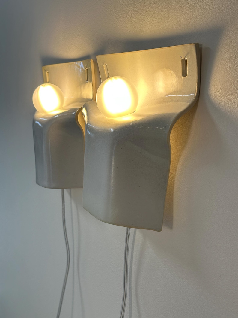 May include: A pair of off-white ceramic wall sconces with a curved, sculptural silhouette. Each fixture features a single glowing spherical light bulb positioned in a recessed top section, with a thin power cord extending downward from the base.