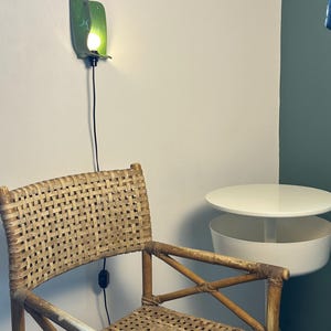 Pu&ograve; includere: Una sedia in rattan intrecciato con finitura marrone chiaro, posizionata accanto a un tavolino circolare bianco con un vano portaoggetti inferiore. Un'applique da parete in ceramica verde con una lampadina accesa &egrave; montata sopra la sedia.