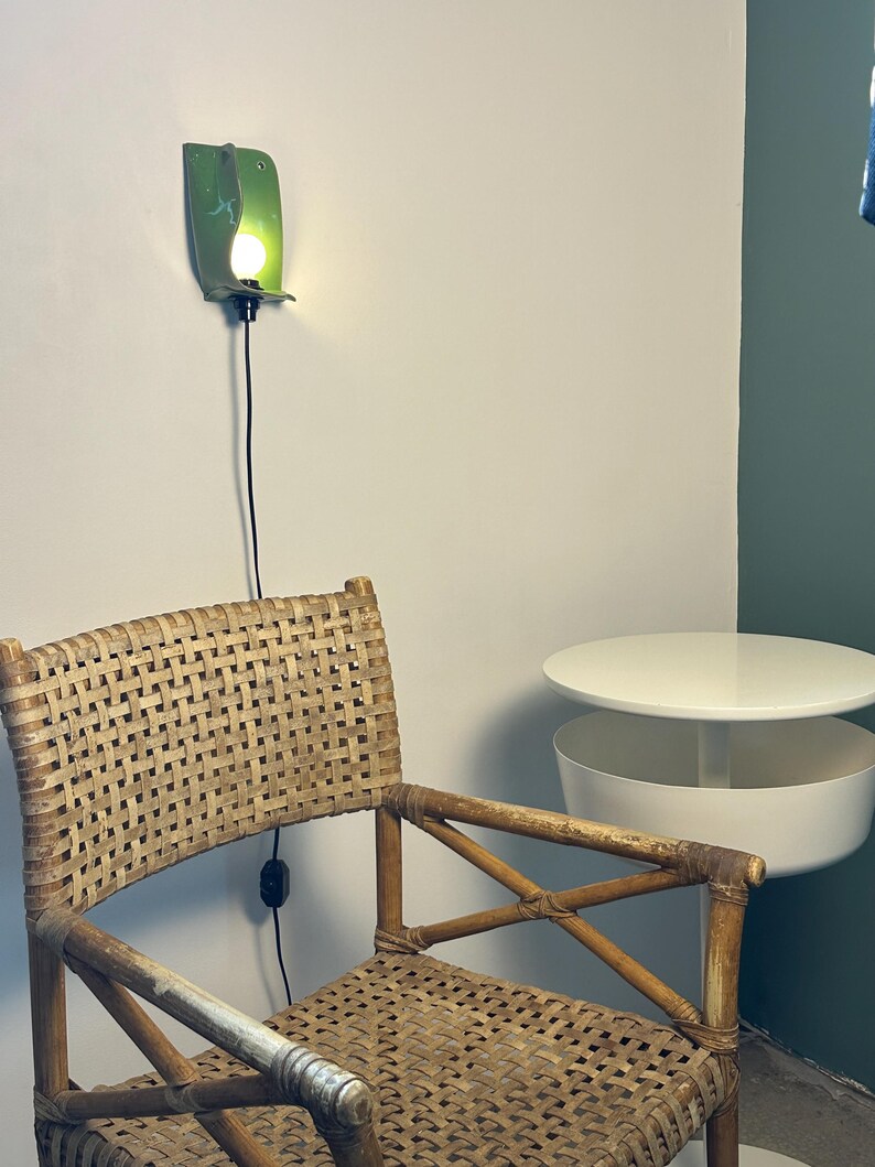 Pu&ograve; includere: Una sedia in rattan intrecciato con finitura marrone chiaro, posizionata accanto a un tavolino circolare bianco con un vano portaoggetti inferiore. Un'applique da parete in ceramica verde con una lampadina accesa &egrave; montata sopra la sedia.