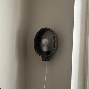 May include: A wall-mounted light fixture with a dark, textured, oval-shaped frame. A white light bulb is centered within the frame. The fixture is mounted on a neutral-colored wall, with a white cord extending downwards.