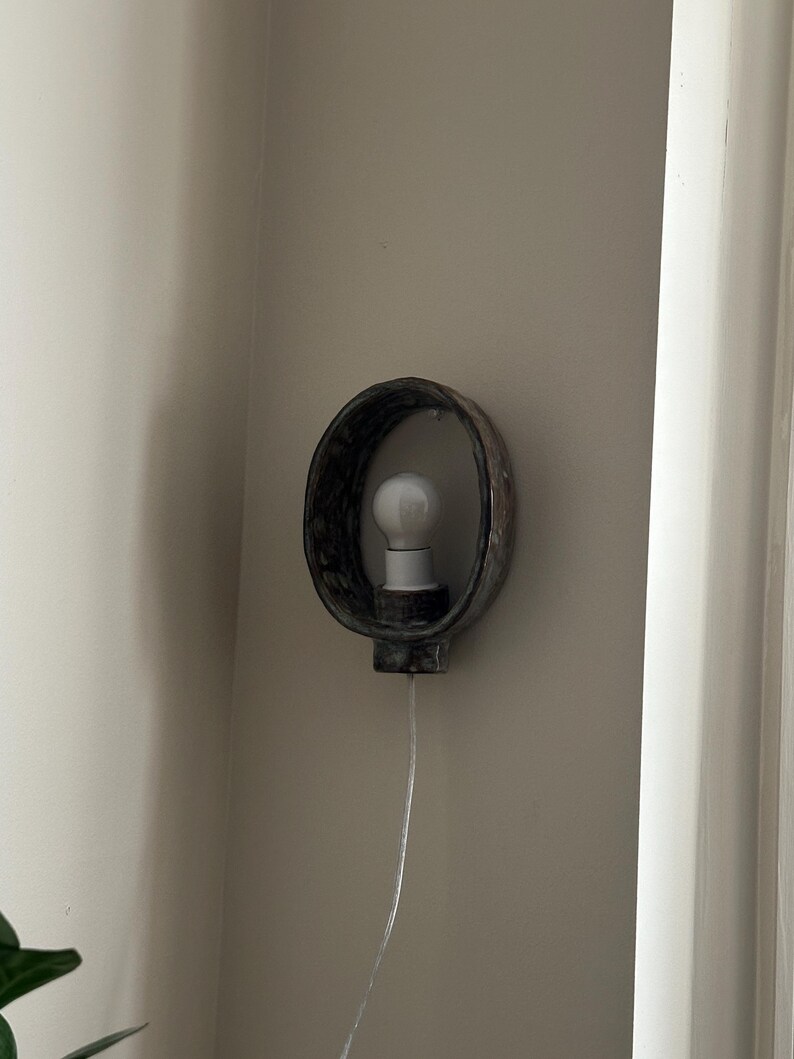 May include: A wall-mounted light fixture with a dark, textured, oval-shaped frame. A white light bulb is centered within the frame. The fixture is mounted on a neutral-colored wall, with a white cord extending downwards.