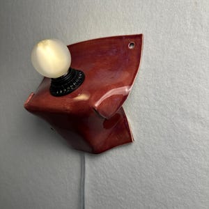 Puede incluir: Un aplique de pared con una base de cer&aacute;mica de color rojo oscuro brillante y forma curva y plegada. Un casquillo negro sostiene una bombilla redonda de color blanco esmerilado. Un cable de alimentaci&oacute;n gris cuelga de la base de cer&aacute;mica.