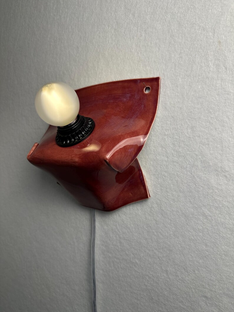 Puede incluir: Un aplique de pared con una base de cer&aacute;mica de color rojo oscuro brillante y forma curva y plegada. Un casquillo negro sostiene una bombilla redonda de color blanco esmerilado. Un cable de alimentaci&oacute;n gris cuelga de la base de cer&aacute;mica.
