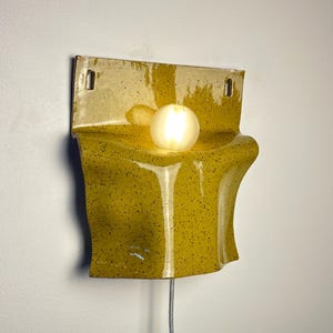 May include: A wall-mounted ceramic sconce featuring a glossy mustard yellow glaze with dark speckles. The fixture has a flat top section with two mounting holes and a curved, undulating lower body that holds a single glowing spherical light bulb.