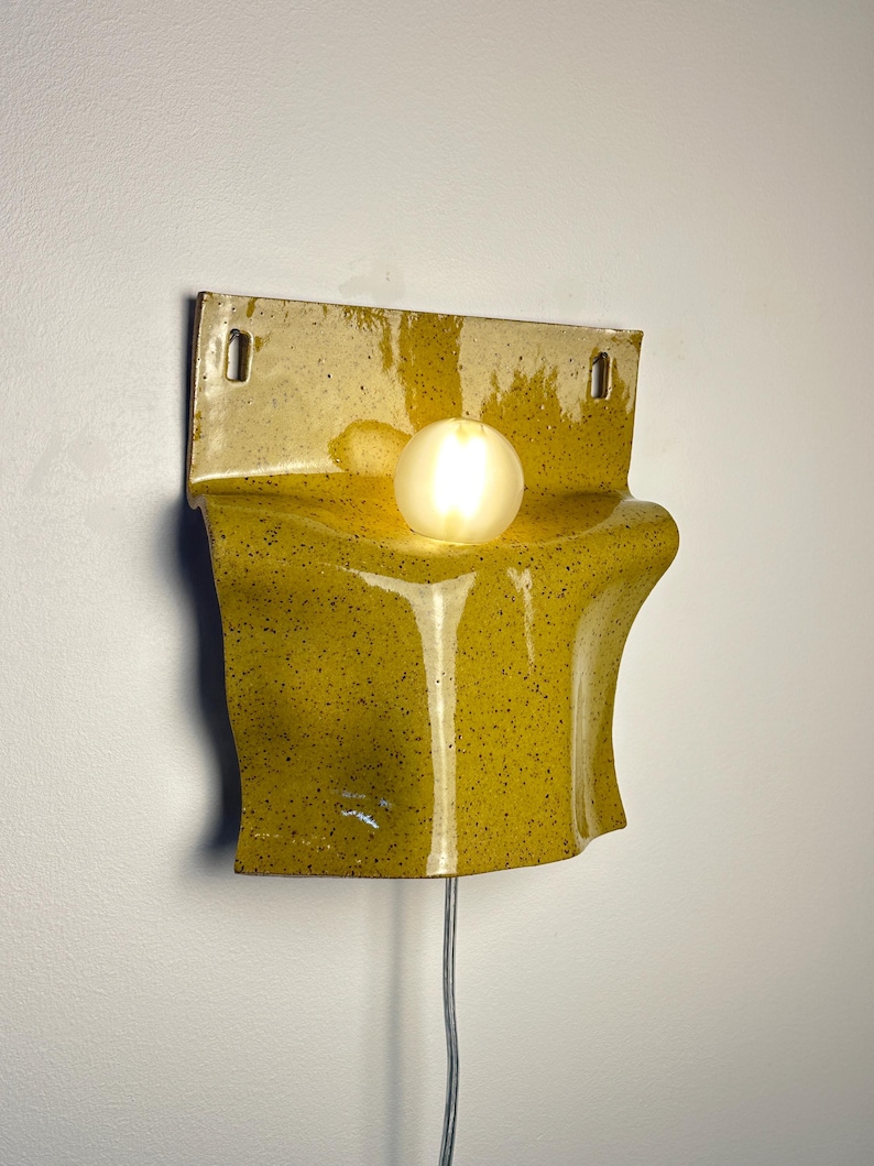 May include: A wall-mounted ceramic sconce featuring a glossy mustard yellow glaze with dark speckles. The fixture has a flat top section with two mounting holes and a curved, undulating lower body that holds a single glowing spherical light bulb.