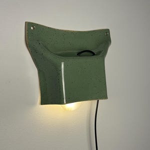 May include: A sage green ceramic wall sconce with a speckled finish and a curved, flared shape. The fixture features a central opening for a light bulb, a black power cord extending from the top, and two mounting holes at the upper corners.
