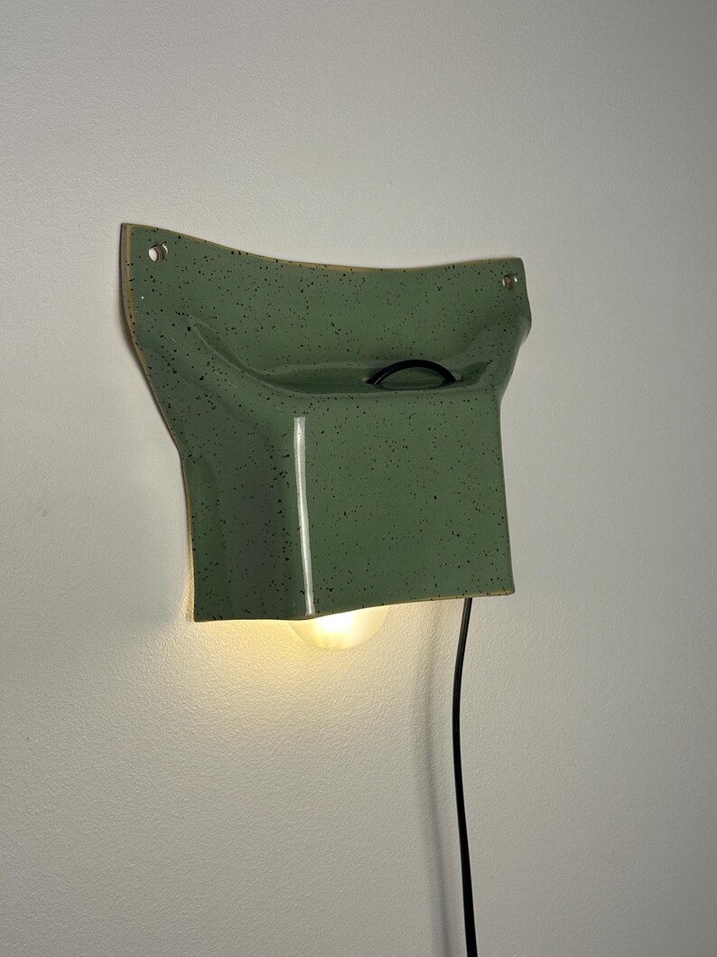 May include: A sage green ceramic wall sconce with a speckled finish and a curved, flared shape. The fixture features a central opening for a light bulb, a black power cord extending from the top, and two mounting holes at the upper corners.