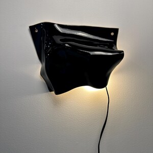 Puede incluir: Una l&aacute;mpara de pared negra brillante con una forma irregular, org&aacute;nica y ondulada. El aplique cuenta con dos peque&ntilde;os orificios circulares de montaje en las esquinas superiores y emite un brillo c&aacute;lido y suave desde su parte inferior, conectado por un cable negro fino.