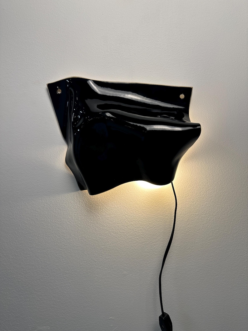 Puede incluir: Una l&aacute;mpara de pared negra brillante con una forma irregular, org&aacute;nica y ondulada. El aplique cuenta con dos peque&ntilde;os orificios circulares de montaje en las esquinas superiores y emite un brillo c&aacute;lido y suave desde su parte inferior, conectado por un cable negro fino.