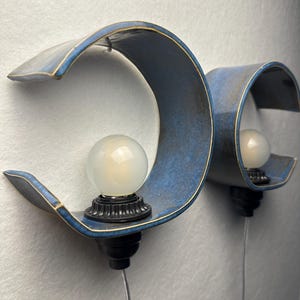 May include: Two wall-mounted sconces featuring curved, blue ceramic shades with gold-toned edges. Each fixture includes a black socket base holding a round, frosted white light bulb. The lamps are positioned side-by-side, each with a thin power cord.