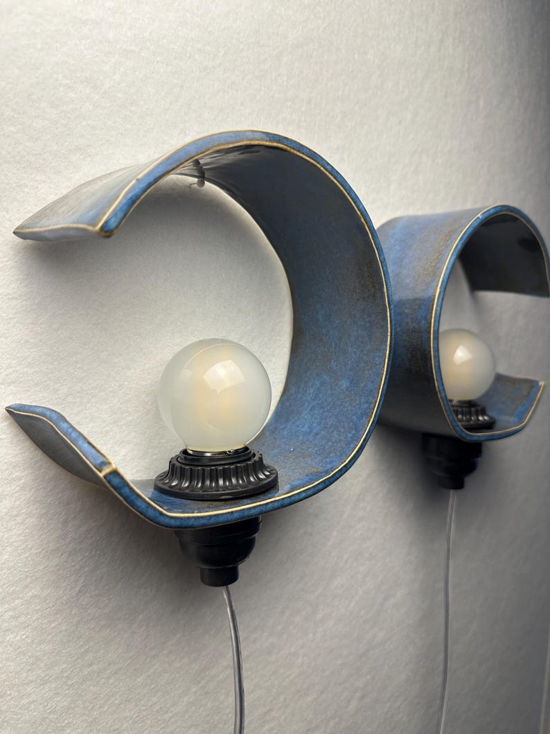 May include: Two wall-mounted sconces featuring curved, blue ceramic shades with gold-toned edges. Each fixture includes a black socket base holding a round, frosted white light bulb. The lamps are positioned side-by-side, each with a thin power cord.