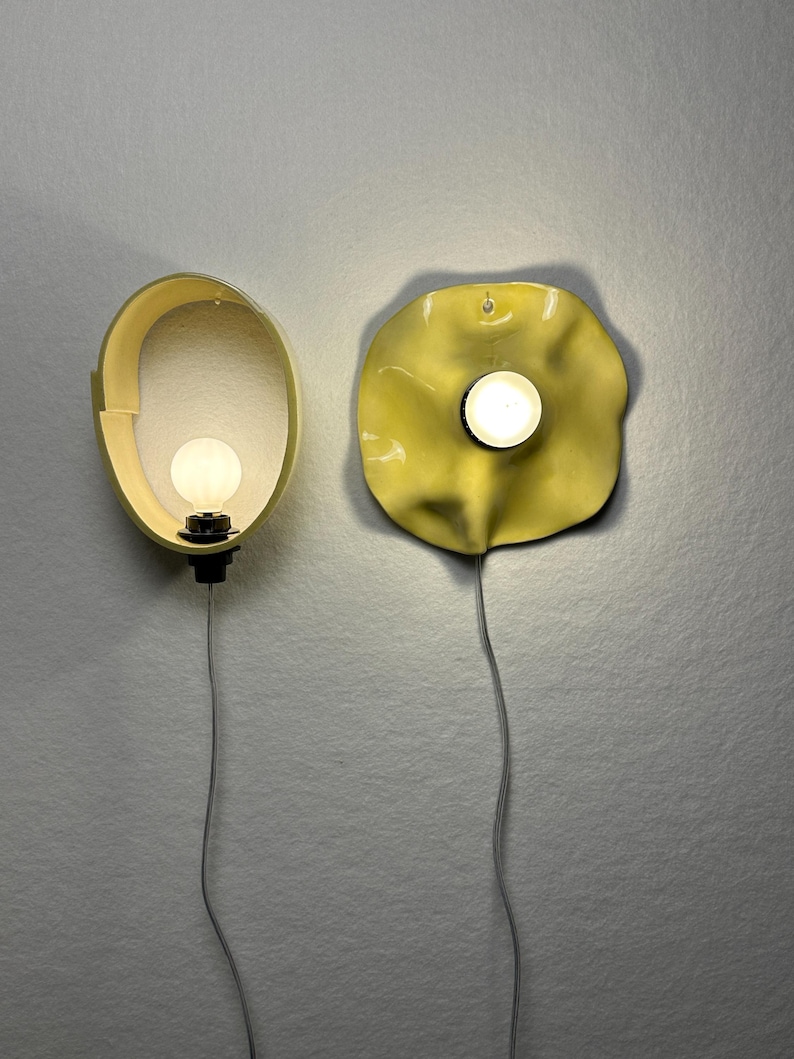 May include: Two wall-mounted light fixtures with yellow shades. The left fixture has an oval frame, while the right fixture features a wavy, organic-shaped disc. Each lamp includes a central glowing white bulb and a thin cord extending downward.