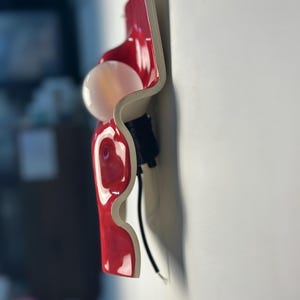 May include: A wall-mounted light fixture featuring a glossy, wavy red ceramic or plastic panel with a white underside. A round, frosted white light bulb is attached to the center of the red surface, supported by a black socket and a black power cord.