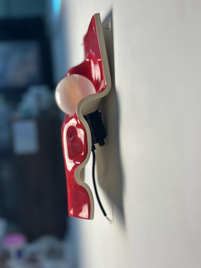 May include: A wall-mounted light fixture featuring a glossy, wavy red ceramic or plastic panel with a white underside. A round, frosted white light bulb is attached to the center of the red surface, supported by a black socket and a black power cord.