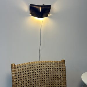 May include: A dark, curved wall-mounted sconce emits a warm, glowing light from its top and bottom edges. A thin, light-coloured power cord hangs vertically from the fixture, which is positioned above a chair with a woven, light-brown cane backrest.