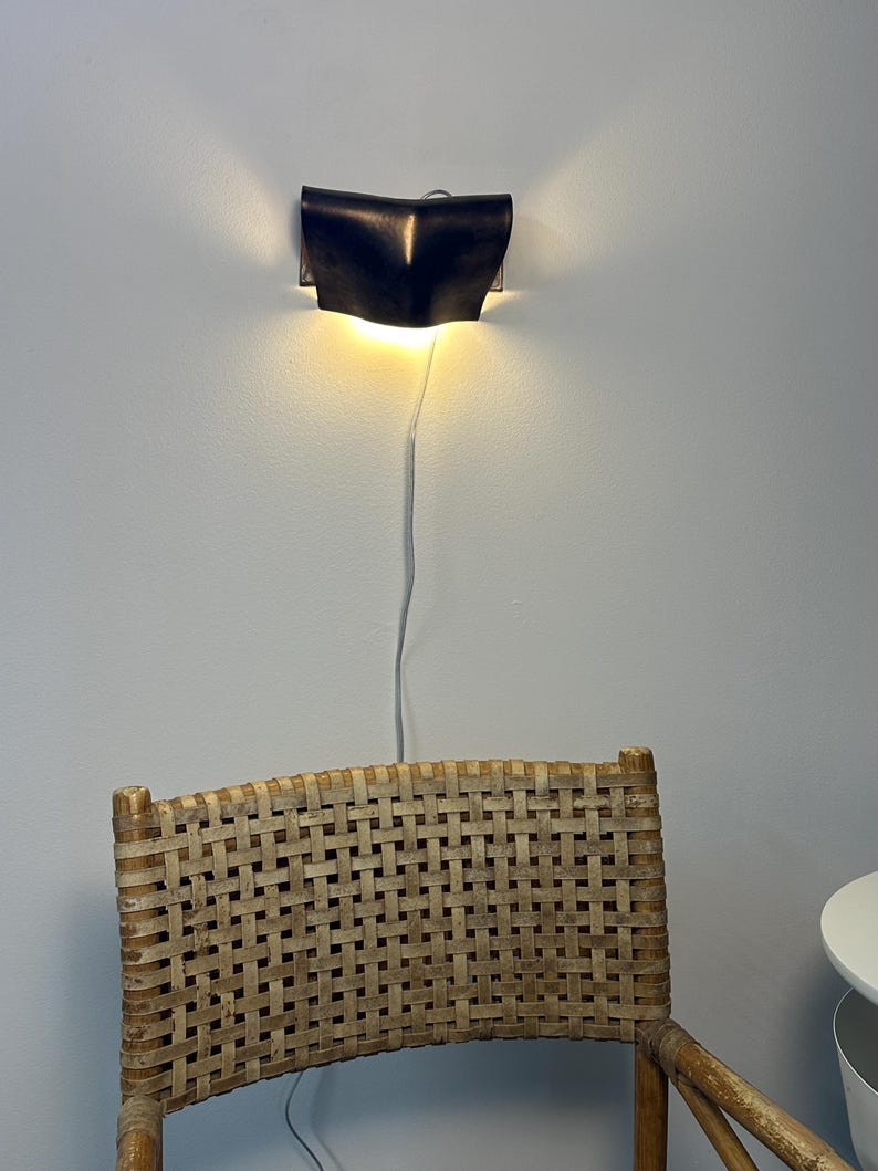 May include: A dark, curved wall-mounted sconce emits a warm, glowing light from its top and bottom edges. A thin, light-coloured power cord hangs vertically from the fixture, which is positioned above a chair with a woven, light-brown cane backrest.