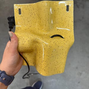 K&ouml;nnte beinhalten: Eine leuchtend gelbe Wandleuchte aus Keramik mit schwarzem Sprenkelmuster. Die Leuchte hat eine geschwungene, organische Form mit zwei rechteckigen Befestigungsl&ouml;chern oben und einem schwarzen Stromkabel, das durch eine mittlere &Ouml;ffnung f&uuml;hrt.