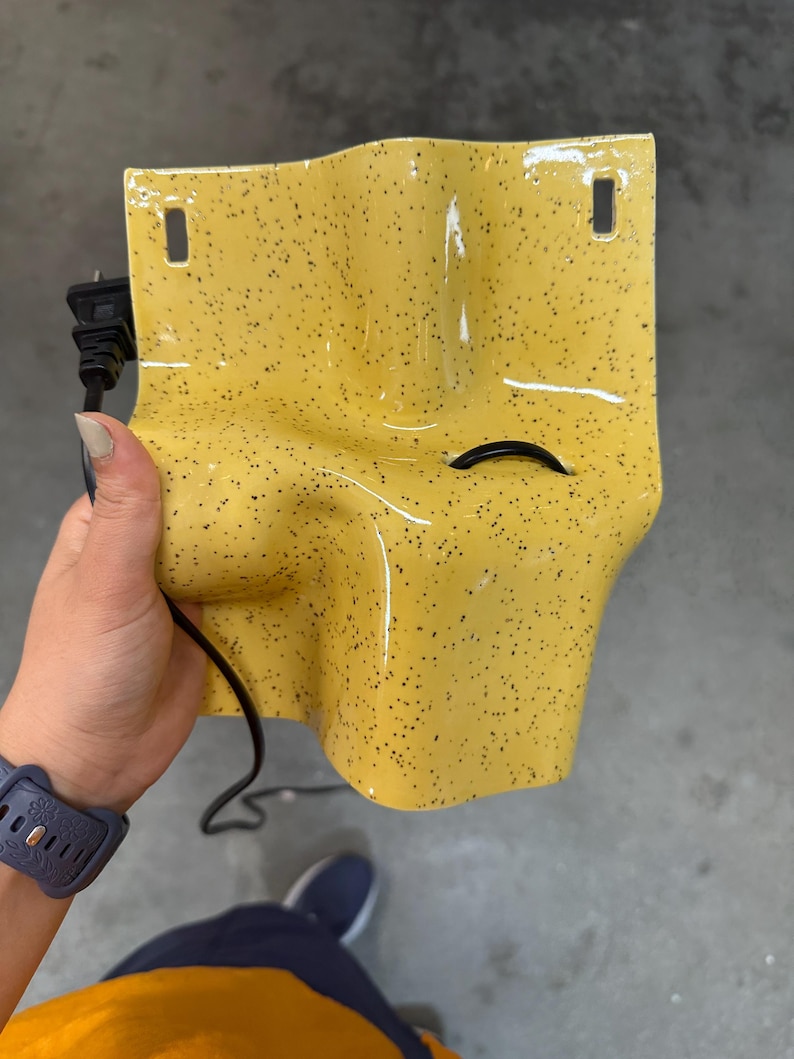 K&ouml;nnte beinhalten: Eine leuchtend gelbe Wandleuchte aus Keramik mit schwarzem Sprenkelmuster. Die Leuchte hat eine geschwungene, organische Form mit zwei rechteckigen Befestigungsl&ouml;chern oben und einem schwarzen Stromkabel, das durch eine mittlere &Ouml;ffnung f&uuml;hrt.