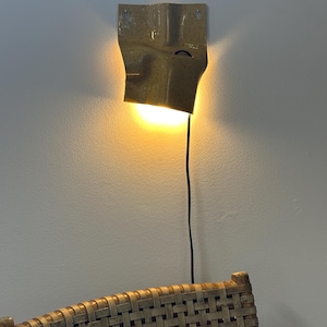 K&ouml;nnte beinhalten: Eine Wandleuchte aus Keramik mit gesprenkelter beiger Glasur und gefalteter, organischer Form. Die Leuchte strahlt ein warmes, nach unten gerichtetes Licht aus und verf&uuml;gt &uuml;ber ein d&uuml;nnes schwarzes Stromkabel an der Unterseite.