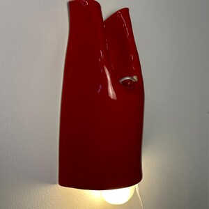 May include: A glossy red ceramic wall sconce with a vertical, folded silhouette. The fixture features a small silver-toned ring detail on the right side and emits a warm, downward-facing light from a bulb positioned at the base of the structure.
