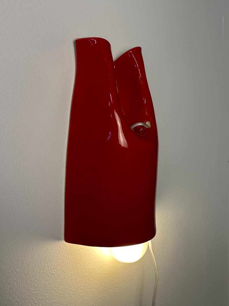 May include: A glossy red ceramic wall sconce with a vertical, folded silhouette. The fixture features a small silver-toned ring detail on the right side and emits a warm, downward-facing light from a bulb positioned at the base of the structure.